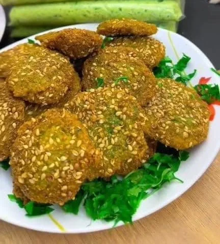 Ta'meya berbalut wijen di piring, dengan kantong-kantong adonan hijau dari kacang fava dan herba di belakangnya