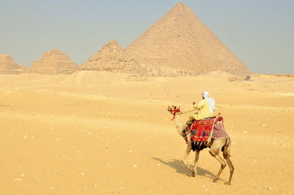 A camel rider crossing the sand in front of a pyramid and its smaller satellite pyramids at Giza